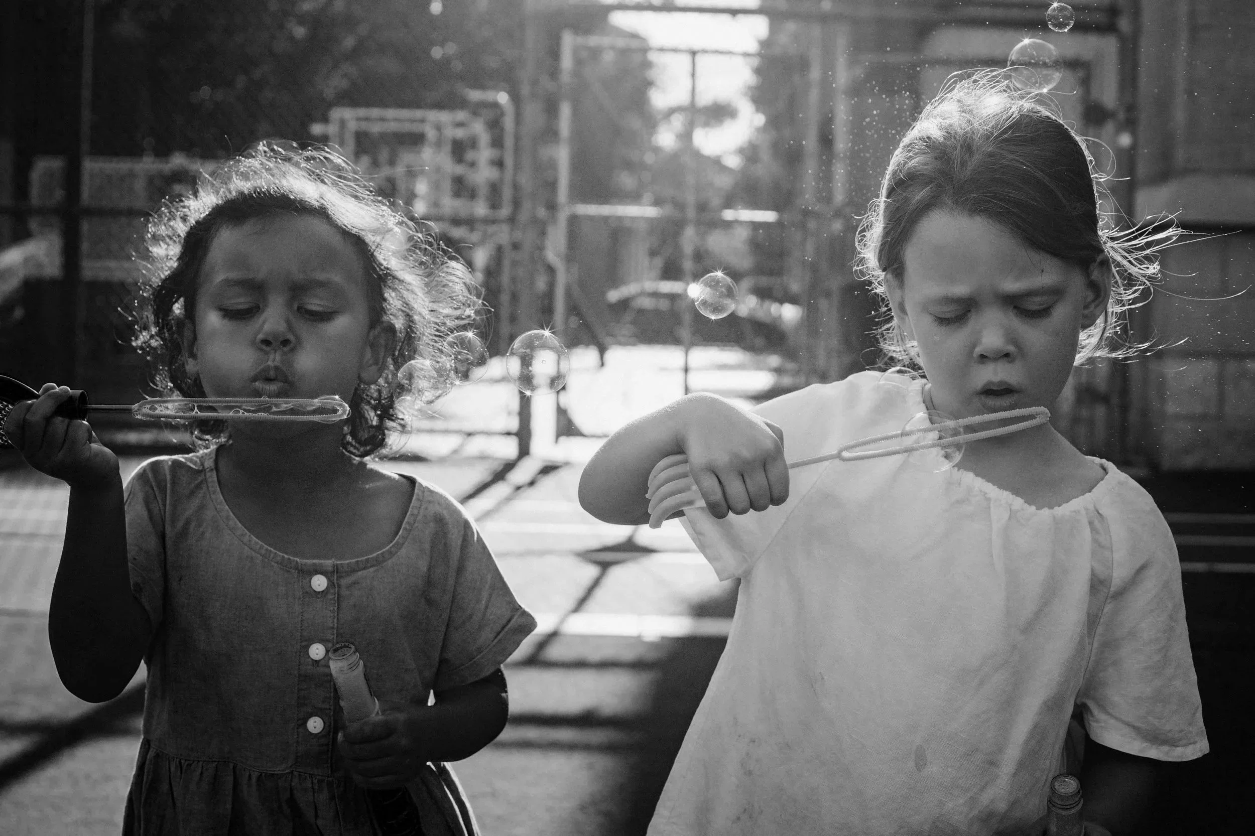 Two young girls blowing soap bubbles outdoors. One girl is on the left, wearing a dress, and the other girl on the right, wearing a T-shirt. Both are focused on the bubbles with sunlight in the background.