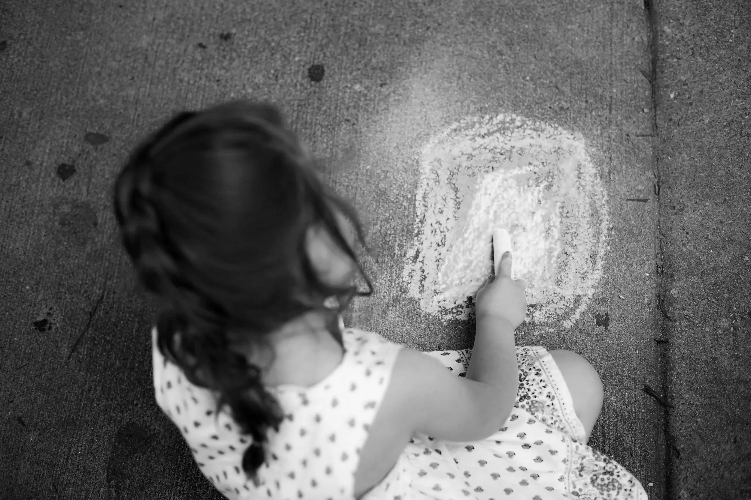 A young girl with long dark hair in a braid, sitting on the ground and drawing with chalk on pavement.