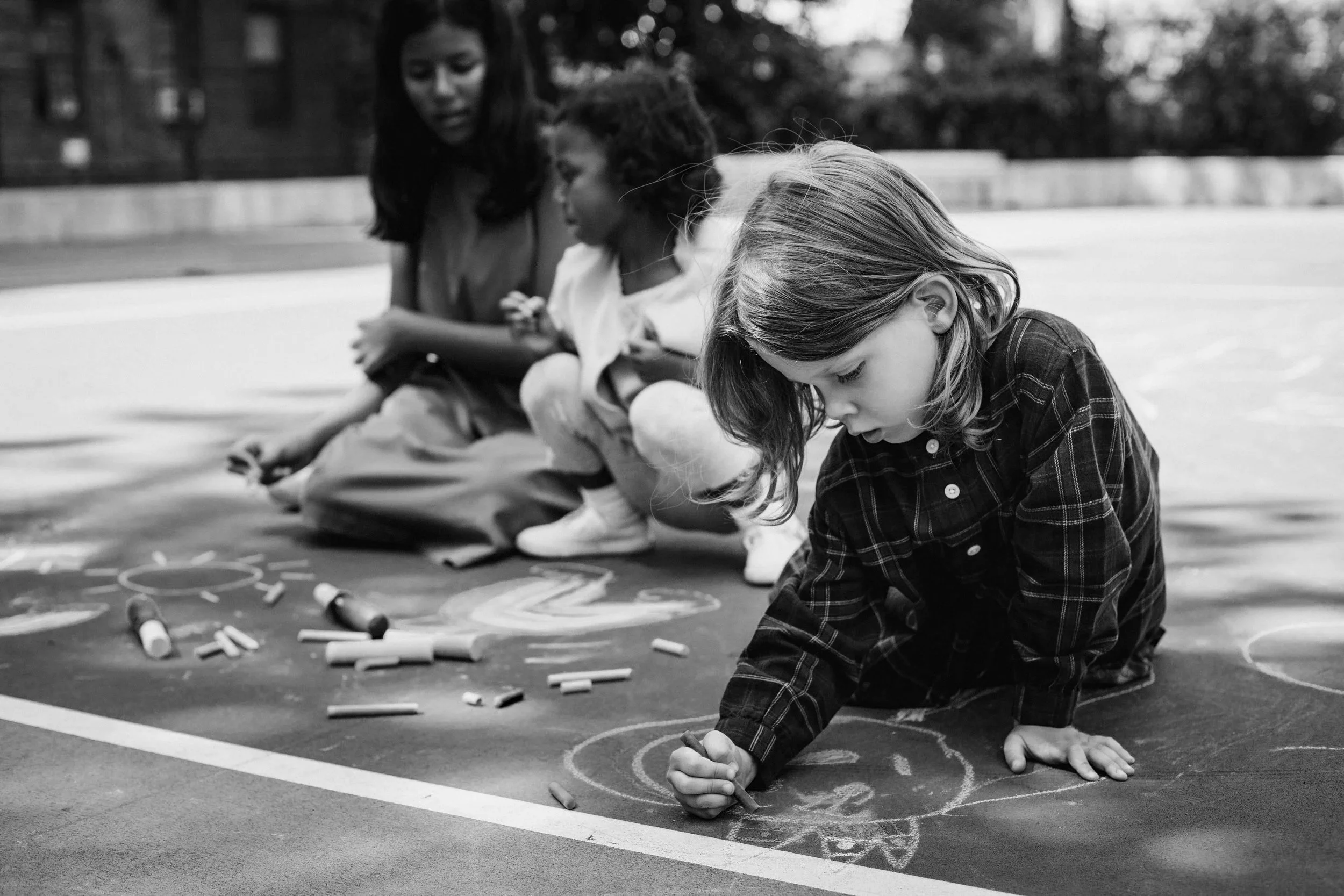 Three young girls drawing with chalk on a basketball court outdoors, with chalk pieces scattered around, and one girl is drawing while kneeling.